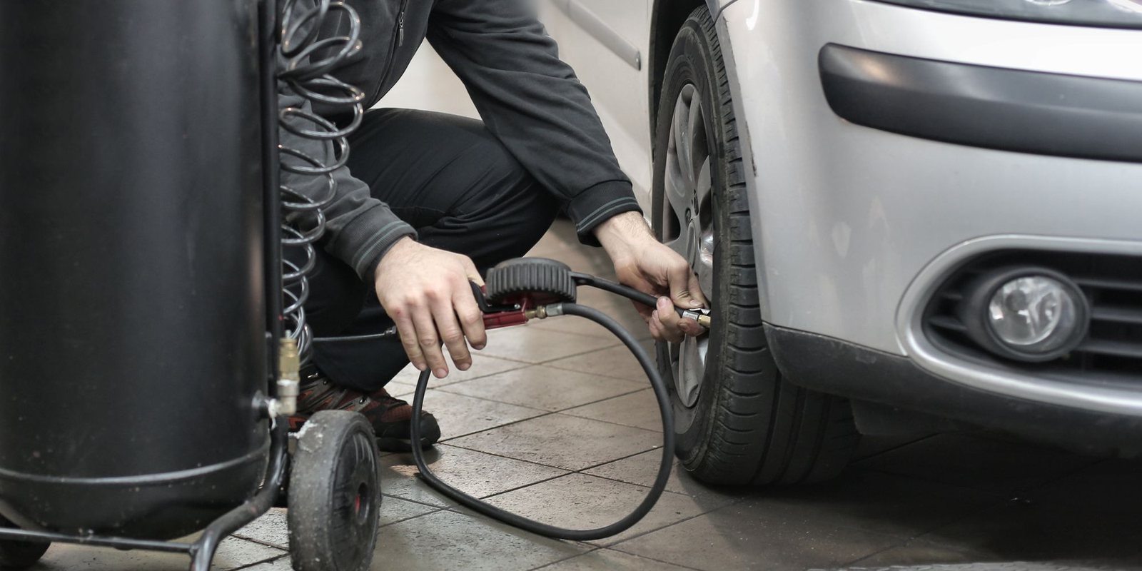 Pessoa agachada ao lado da roda dianteira calibrando o pneu de um carro prata com a mangueira do compressor