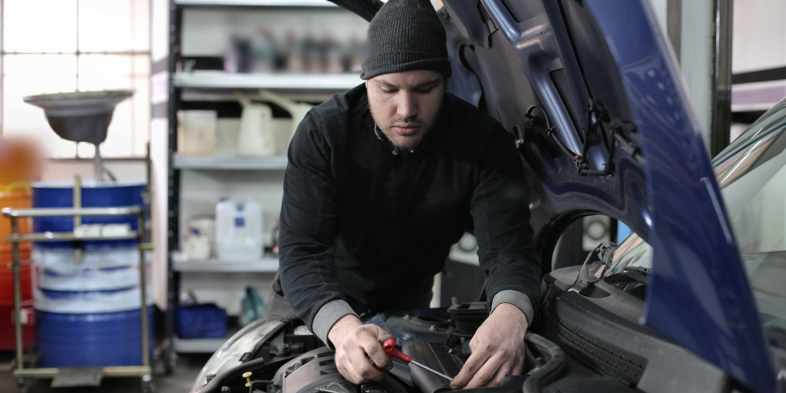 Mecânico de gorro preto ajustando peças no compartimento do motor de um carro azul, com prateleiras de frascos ao fundo