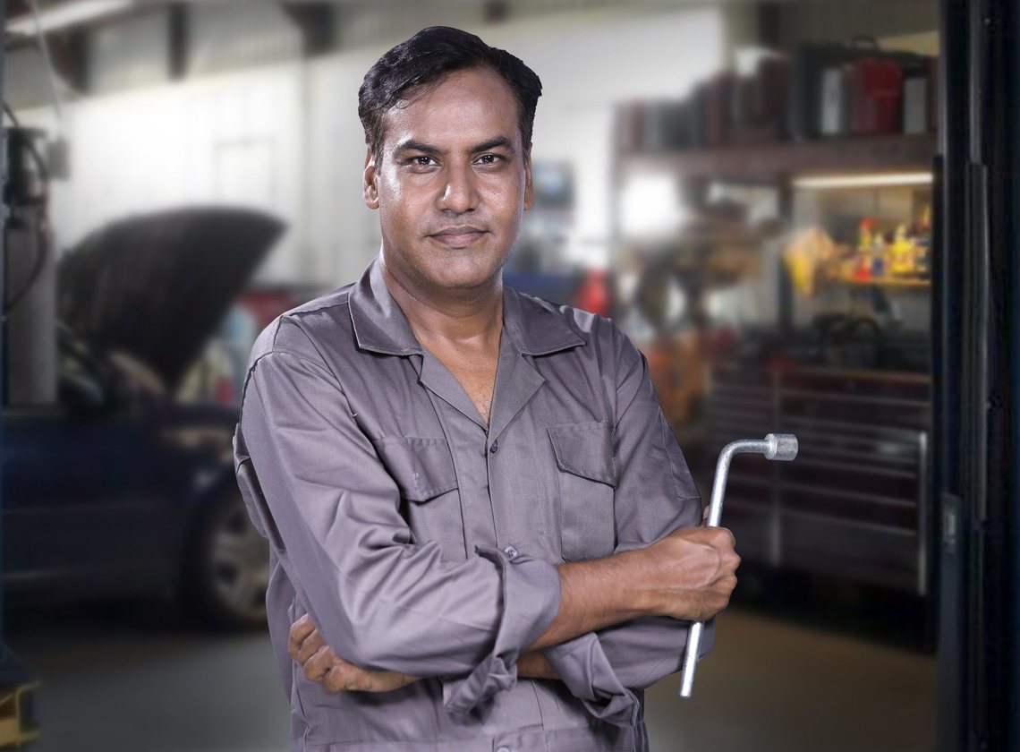 Mecânico de camisa cinza segurando uma chave de roda, em pé numa oficina com um carro de capô aberto ao fundo